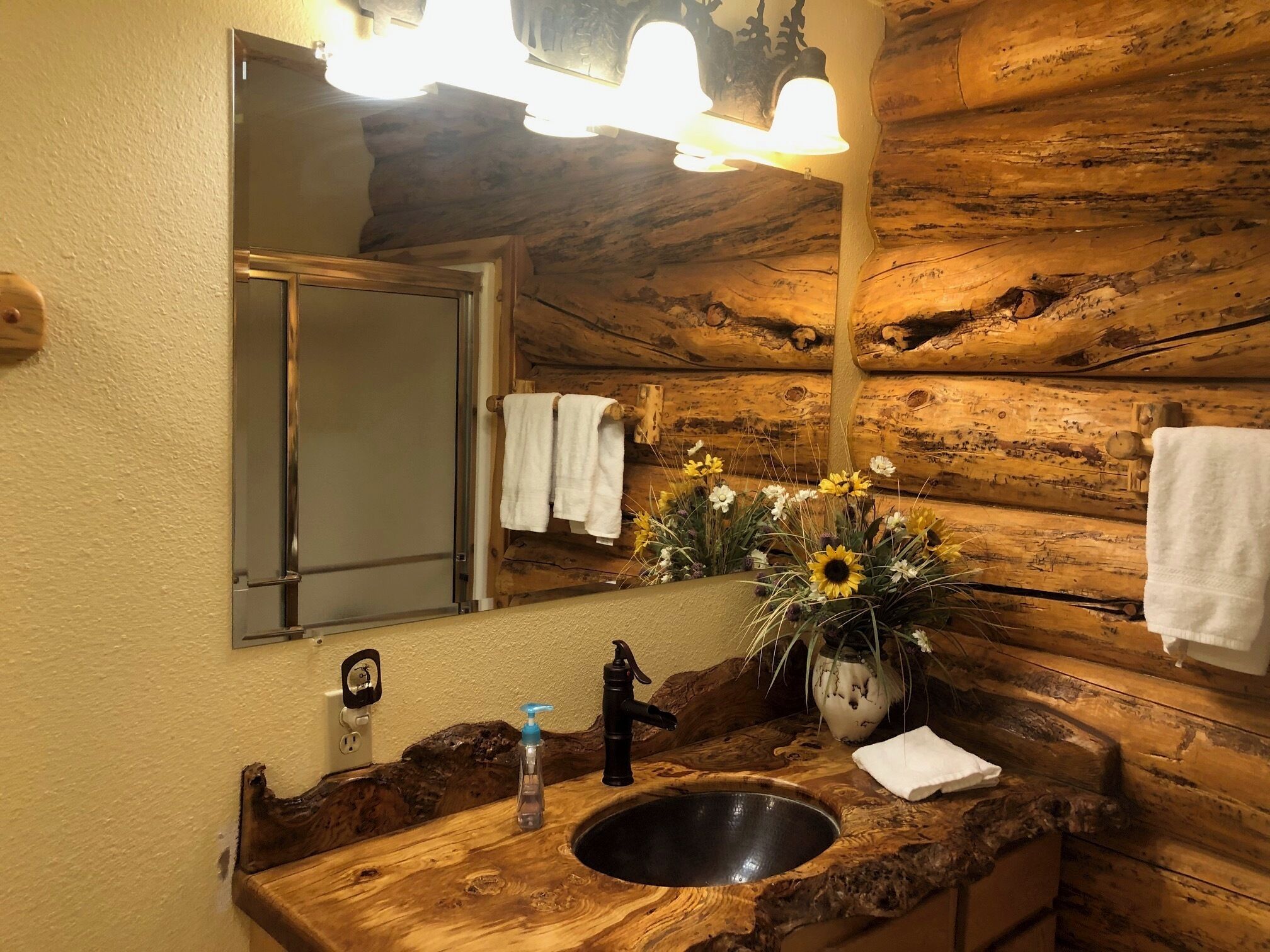 A rustic bathroom with a wooden vanity, a sink, a mirror, and a vase with flowers.