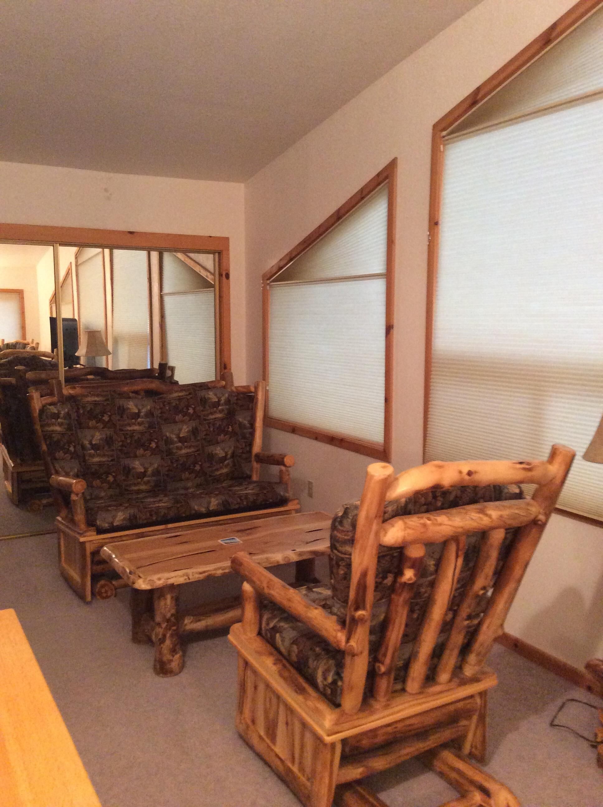 Un salon de style rustique, avec des meubles en bois, notamment un canapé et des fauteuils, et de grandes fenêtres équipées de stores.