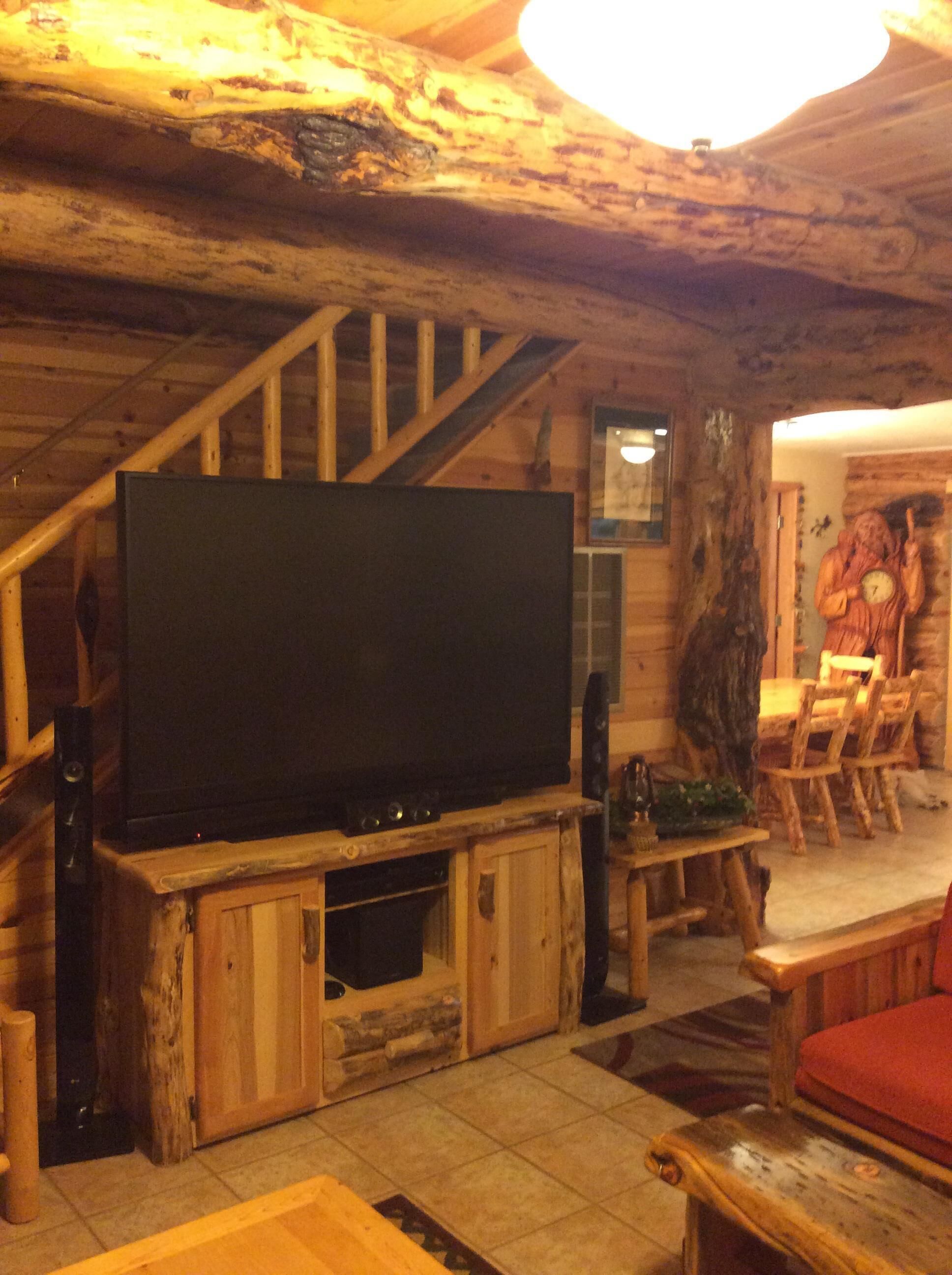 A rustic interior with a wooden staircase, a flat-screen TV on a wooden stand, and a wooden dining table with chairs.