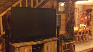A rustic interior with a wooden staircase, a flat-screen TV on a wooden stand, and a wooden dining table with chairs.