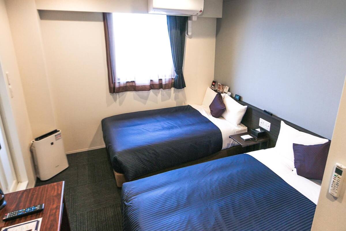A hotel room with two beds, a desk, a chair, an air conditioning unit, and a window with curtains.