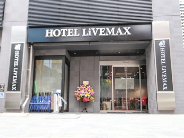 The entrance of HOTEL LIVEMAX with a large glass facade, a flower arrangement outside, and a signboard above the entrance.