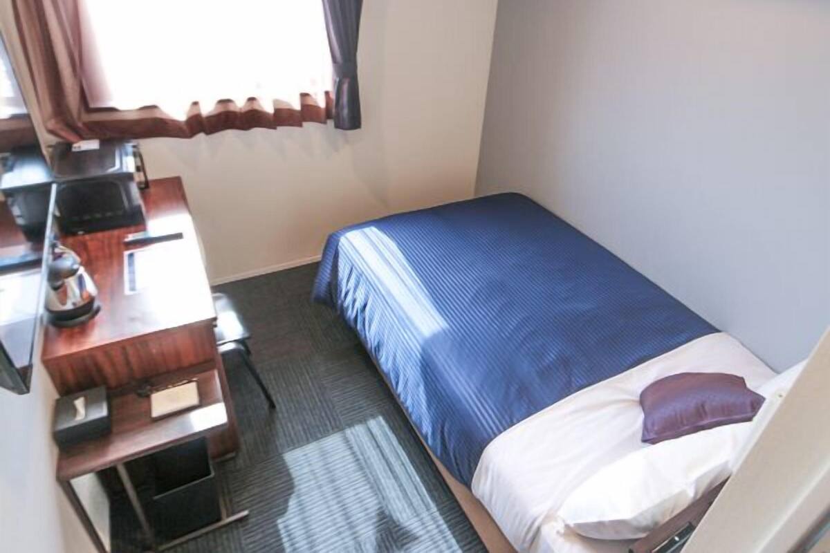 A small, single-bed room with a desk and chair, a window with curtains, and a bed with a blue blanket and pillows.