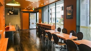 Interior moderno de restaurante con mesas y sillas de madera, iluminación colgante y grandes ventanales con vistas a la calle.