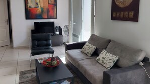 A modern living room with a grey sofa, a black armchair, a coffee table with a fruit bowl, a flat-screen TV, and a painting on the wall.