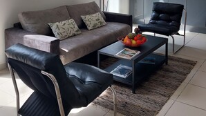 A modern living room with a grey sofa, black leather chair, and a coffee table with a fruit bowl. There is a painting on the wall and a kitchen in the background.