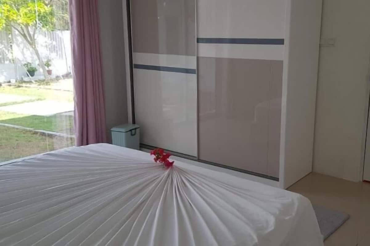 A bedroom with a bed covered in white sheets, a small trash bin, and a door with a smoke alarm.