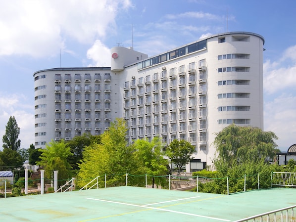 Un hotel de varios pisos con fachada curva, numerosos balcones y una cancha de tenis en primer plano.