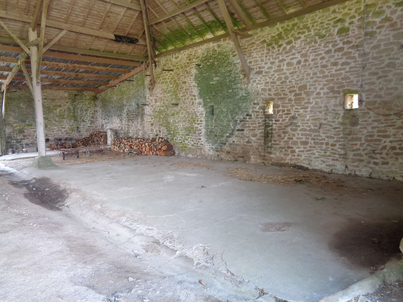 Una habitación con paredes de piedra, suelo de concreto, vigas de madera y una pila de leña.