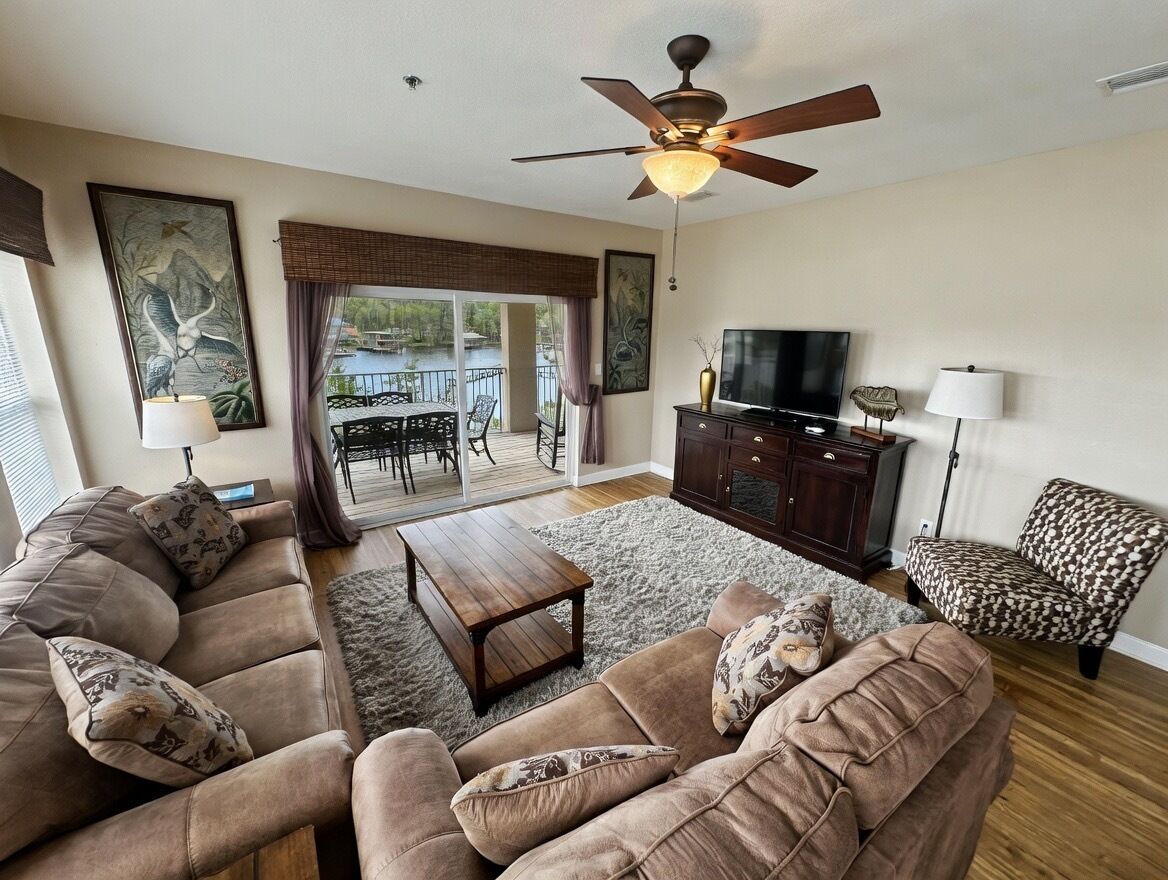 A living room with a sofa set, a television, a ceiling fan, and a view of the outdoors.