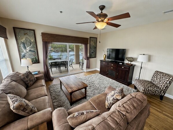 A living room with a sofa set, a television, a ceiling fan, and a view of the outdoors.