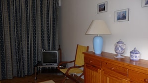 Een woonkamer met een televisie, een lamp, een stoel, een kast en een gordijn met een patroon.