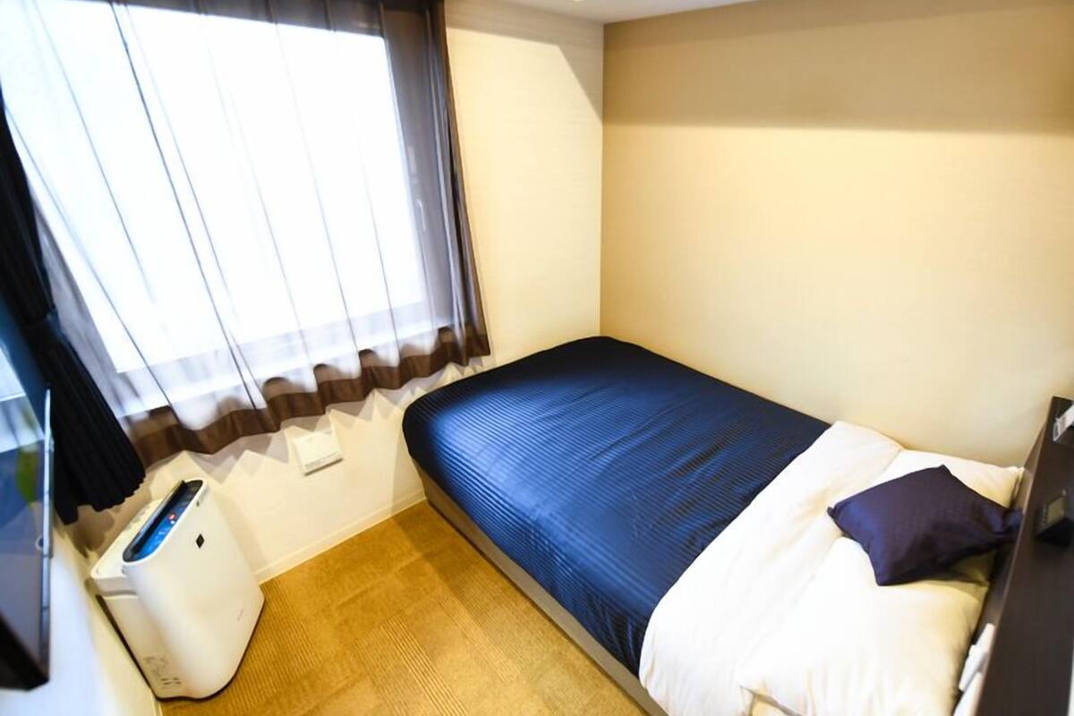 A small, single-bed room with a window, a desk, and a trash bin.