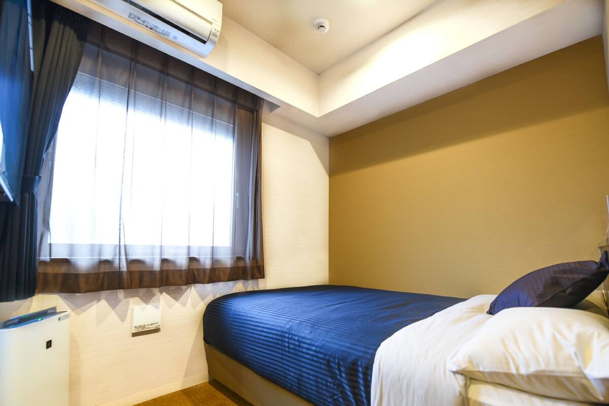 A bedroom with a bed, a window with curtains, an air conditioning unit, and a bedside table.