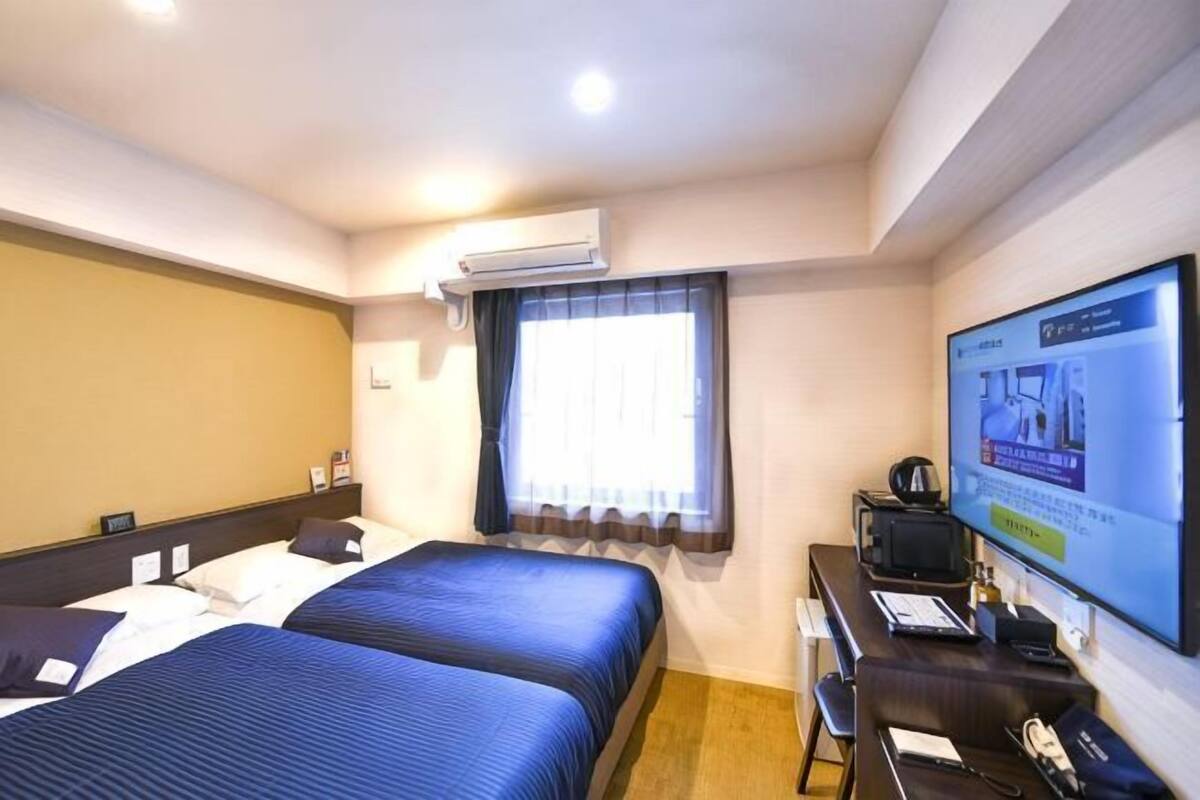 A hotel room with two beds, a desk, a television, and an air conditioning unit.