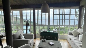 A spacious room with a large window offering a view of the sea and greenery. There is a telescope, a table with a book, and two sofas.