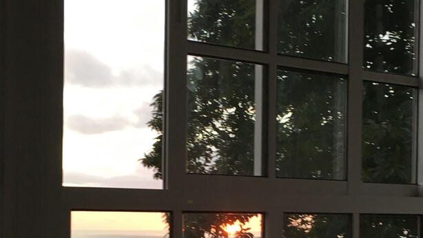 A room with a large window overlooking a sunset and a body of water. There is a table with a tray holding glasses and a bottle.