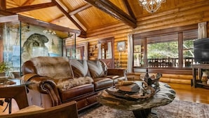 A cozy log cabin interior with a leather sofa, a rustic coffee table, and a large window overlooking a deck. - Cinnamon Valley - The Cattail (Eureka Springs)