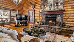 A log cabin interior with a stone fireplace, a mounted deer head, a chandelier, and a television. - Cinnamon Valley - The Cattail (Eureka Springs)