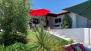 Un espace extérieur moderne, aménagé avec des sièges, un parasol rouge, un mur blanc et un escalier menant à un patio couvert.