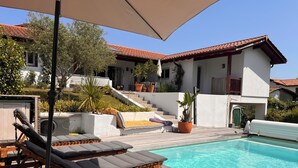Un espace aménagé au bord de la piscine, avec des chaises longues, une terrasse en bois et un bâtiment blanc surmonté d’un toit recouvert de tuiles rouges.