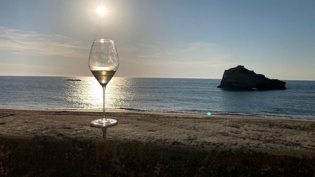 Un verre de vin blanc sur une plage, avec une vue dégagée sur l’océan et une formation rocheuse à l’horizon.