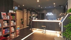 A hotel reception area with a reception desk, a bookshelf with magazines, and a red chair. - Hotel Livemax Shinsaibashi East (Osaka)