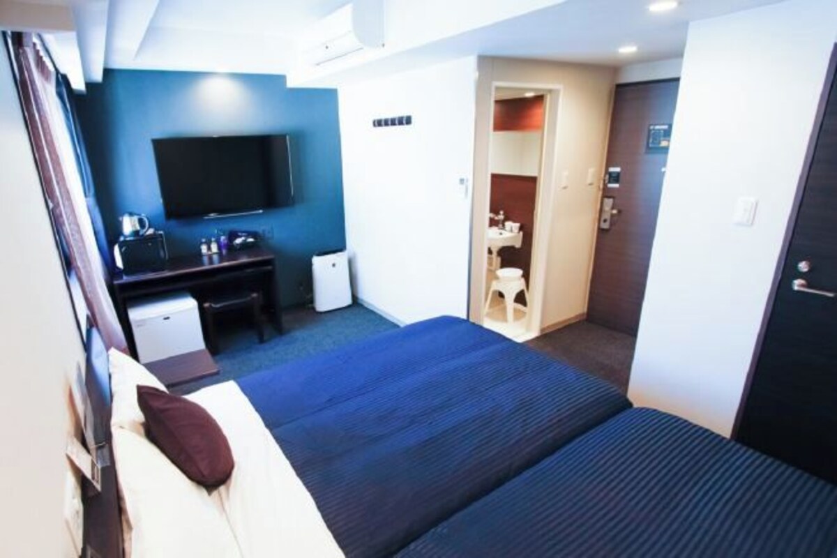 A hotel room with a bed, a desk with a flat-screen TV, a small seating area, and a bathroom visible through an open door.