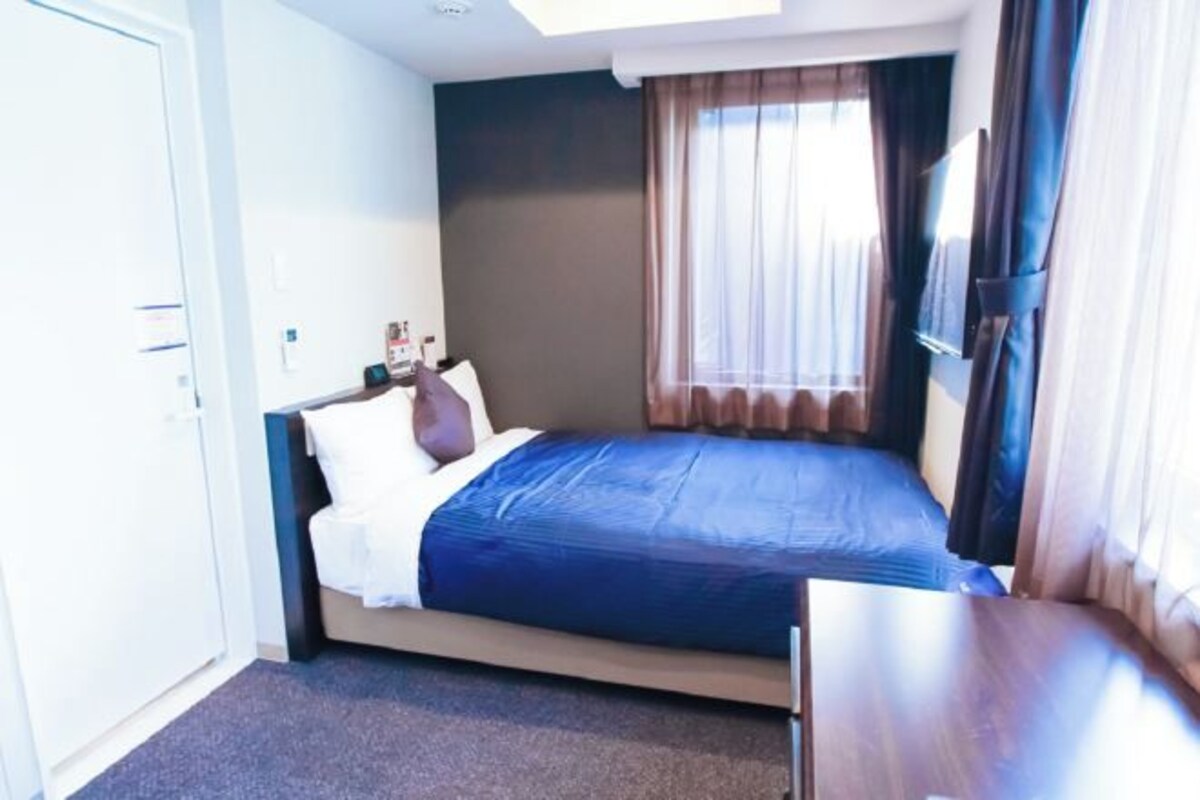 A hotel room with a bed, a desk, a window with curtains, and a door.