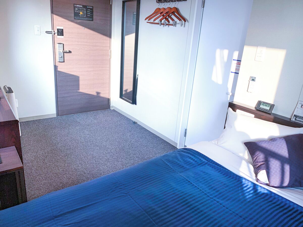 Una habitación de hotel con cama, escritorio, armario, espejo y puerta que da a un pasillo.