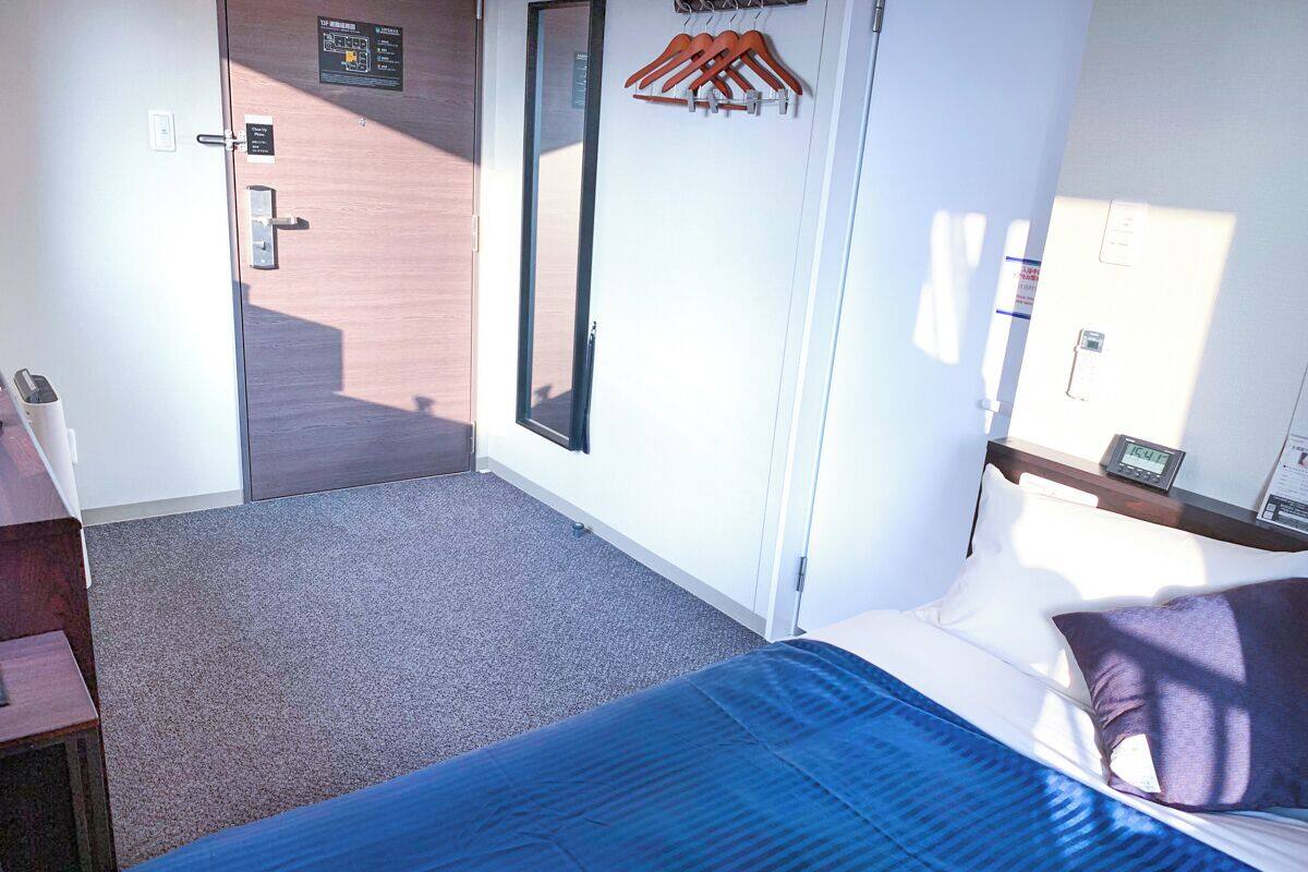 A hotel room with a bed, a desk, a wardrobe, a mirror, and a door leading to a corridor.