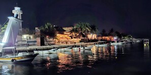 A nighttime harbor scene with boats docked, a lighthouse, and buildings with warm lighting.