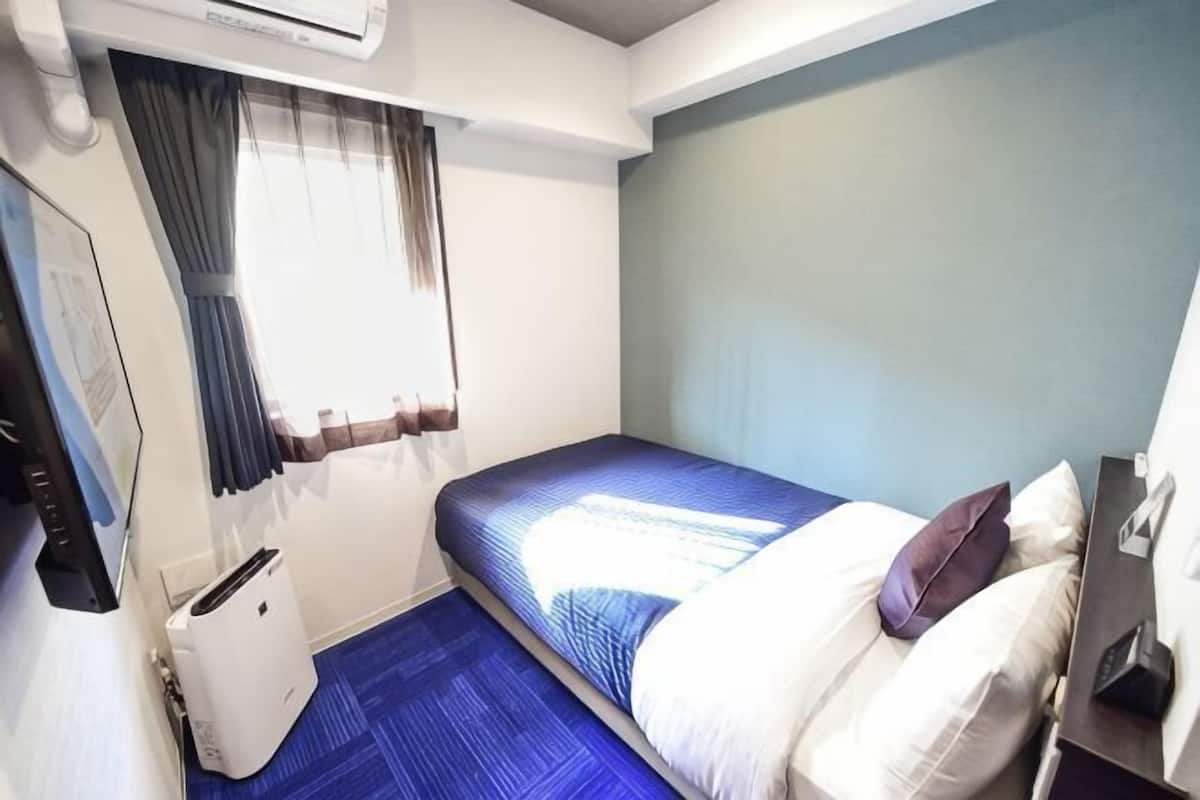 A small, compact room with a single bed, a bedside table, a chair, and a window with curtains.