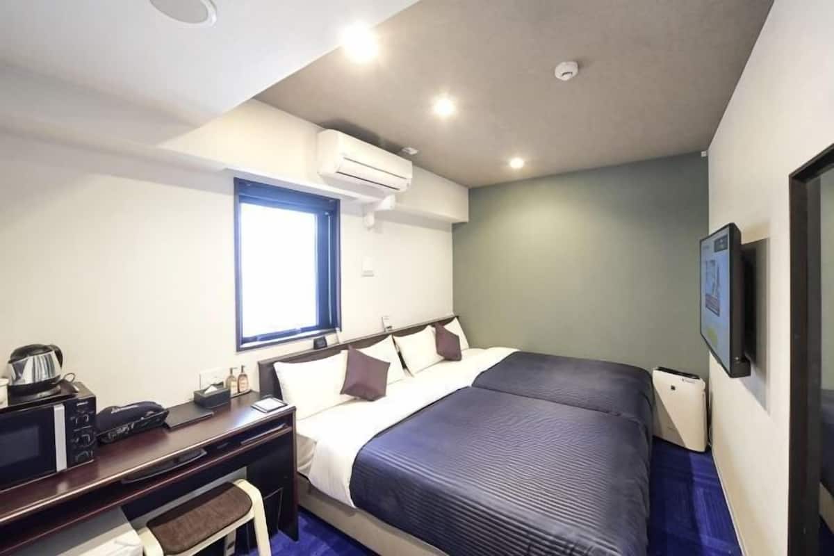 A hotel room with a bed, a desk with a chair, a window, an air conditioning unit, and a television.