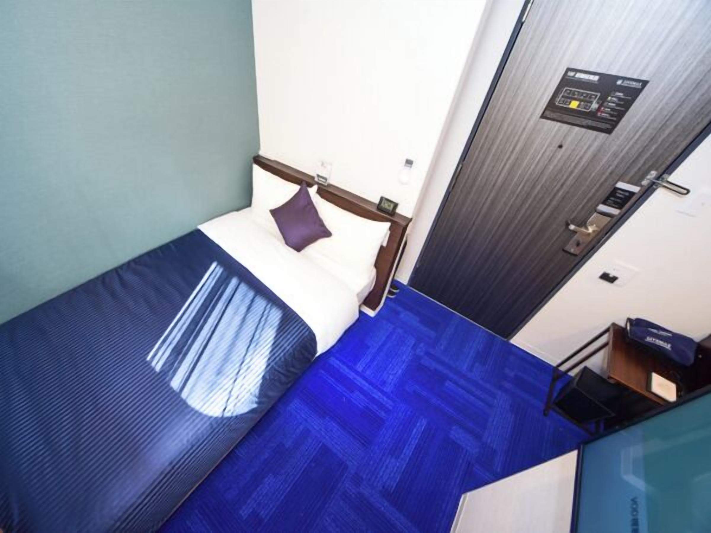 Camera d'albergo compatta con un letto singolo, una scrivania piccola e una porta dotata di pannello di controllo.
