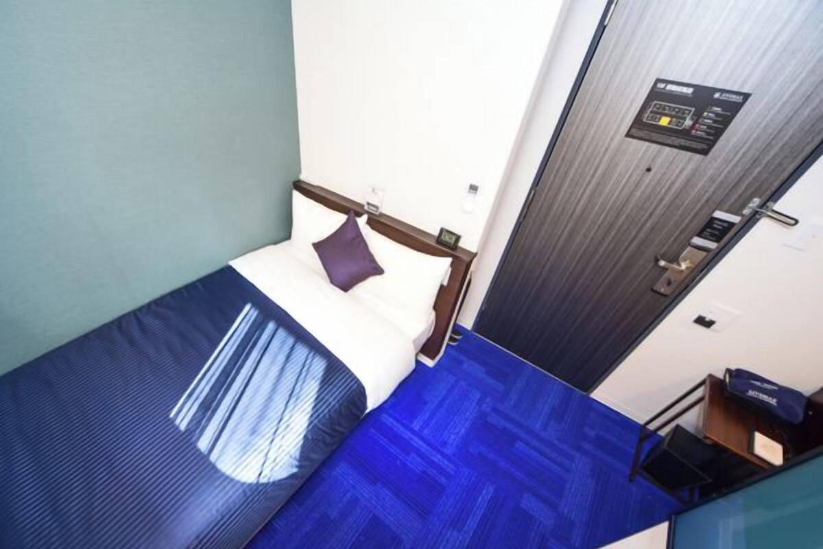 A compact hotel room with a single bed, a small desk, and a door with a control panel.