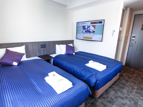 Quarto de hotel com duas camas, cada uma com colcha azul e travesseiros brancos. Há uma mesinha entre as camas e uma televisão fixada na parede.