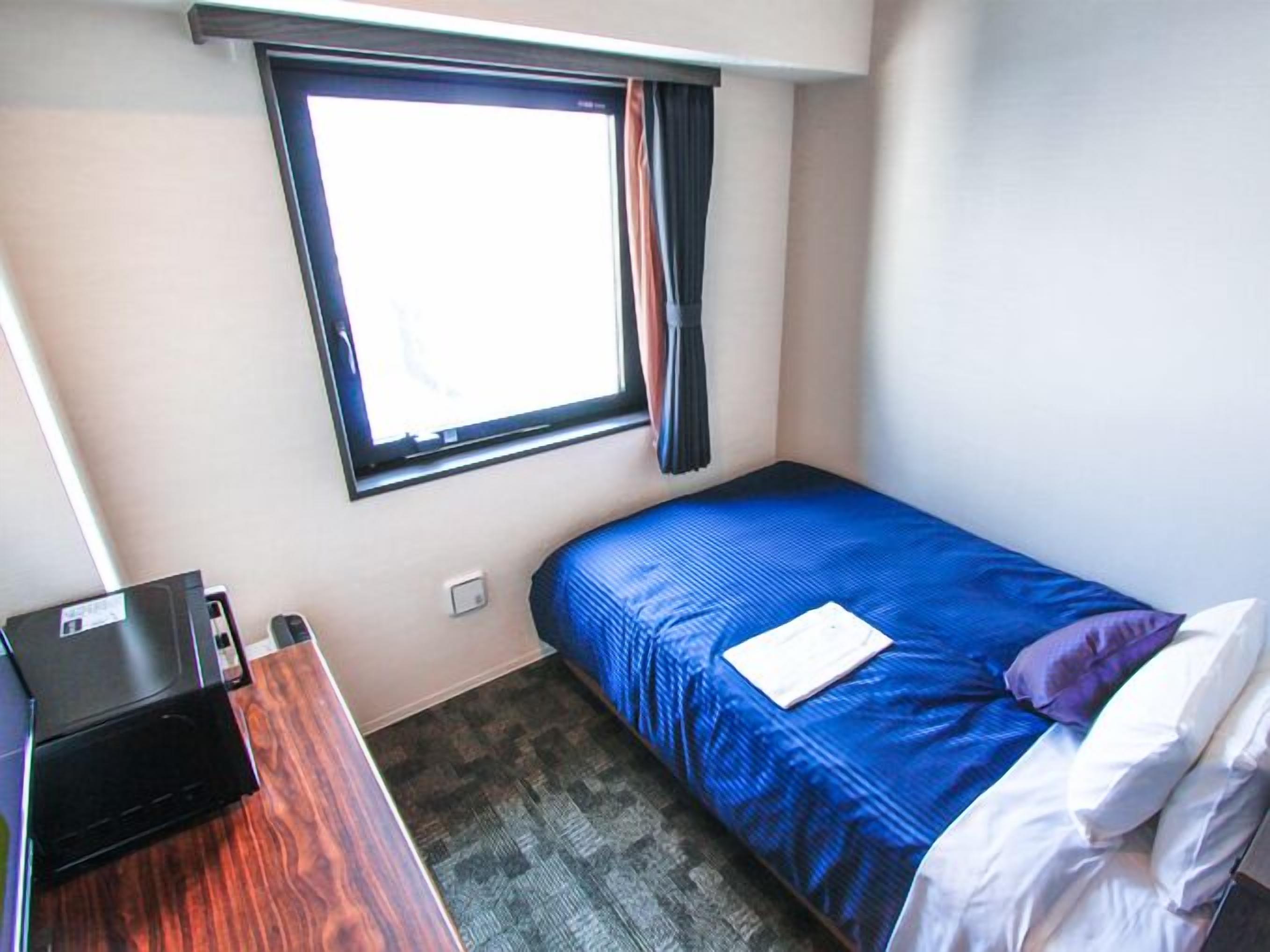 A small, single-bed room with a window, a desk with a printer, and a carpeted floor.