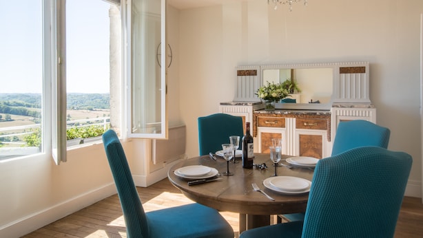 A dining area with a round table, teal chairs, and a view of the countryside through a large window.