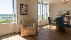 A modern living room with a beige armchair, a dining area with teal chairs, and a view of greenery through large windows.