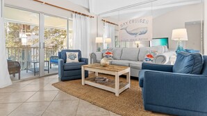 A living room with a sofa, armchairs, a coffee table, and a large window overlooking a balcony. - 2BR Beach Villa w/6-Seat GOLF CART! Near Pool, Tram, Beach. Screened Balcony (Miramar Beach)