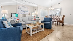 A living room with a grey sofa, blue armchairs, a coffee table, and a dining area with a chair and table. - 2BR Beach Villa w/6-Seat GOLF CART! Near Pool, Tram, Beach. Screened Balcony (Miramar Beach)