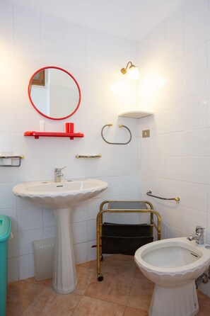 A bathroom with a pedestal sink, a round mirror, a bidet, and a handrail.