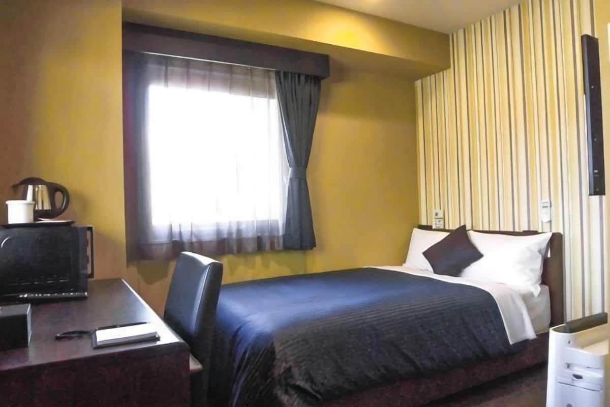 A hotel room with a bed, a desk with a computer, a chair, a window with curtains, and a striped wall.