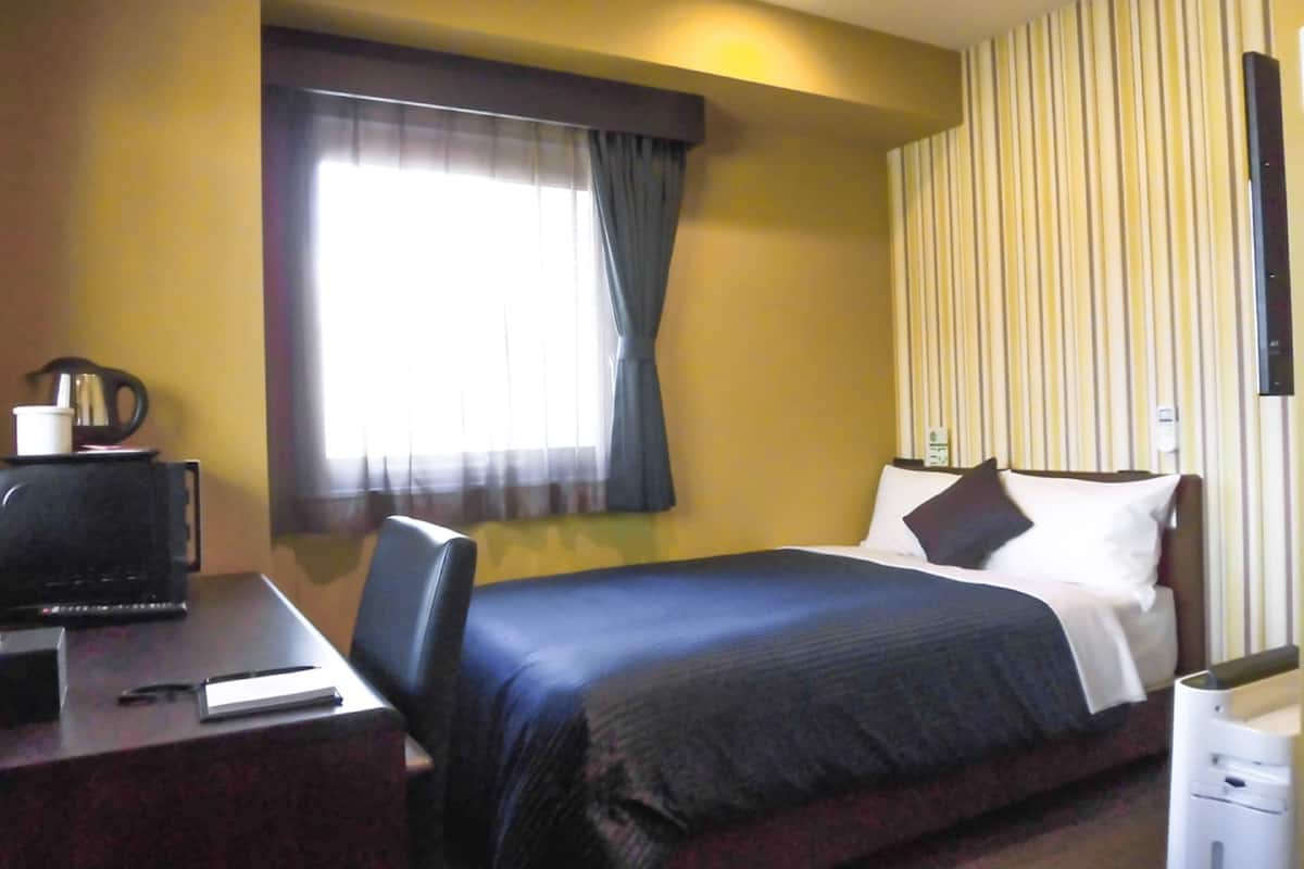 A hotel room with a bed, a desk with a computer, a coffee maker, and a window with curtains.