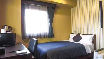 A hotel room with a bed, a desk with a computer, a coffee maker, and a window with curtains.
