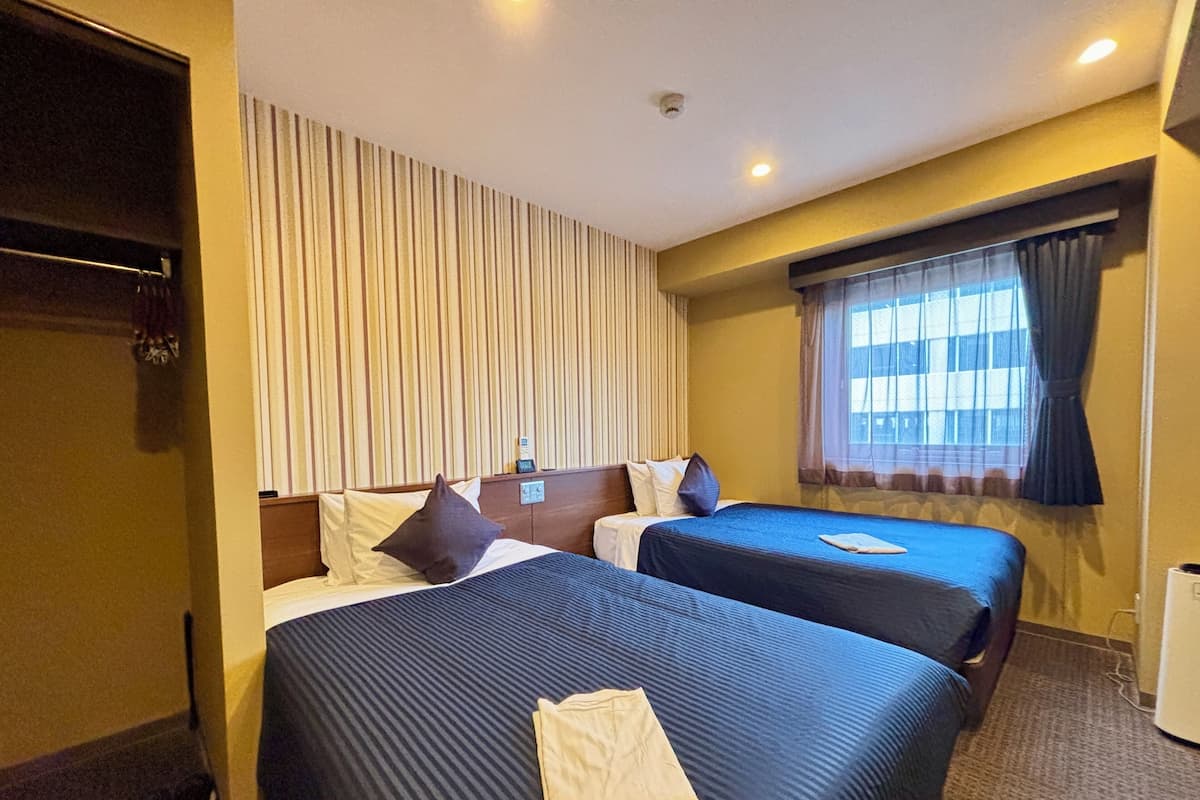A hotel room with two beds, a window with curtains, a wall-mounted shelf, and a small table.