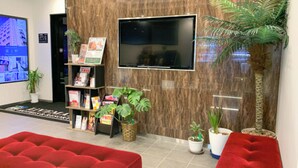 Un hall d’entrée avec un canapé rouge, un téléviseur fixé au mur, une bibliothèque remplie de magazines et des plantes en pot.
