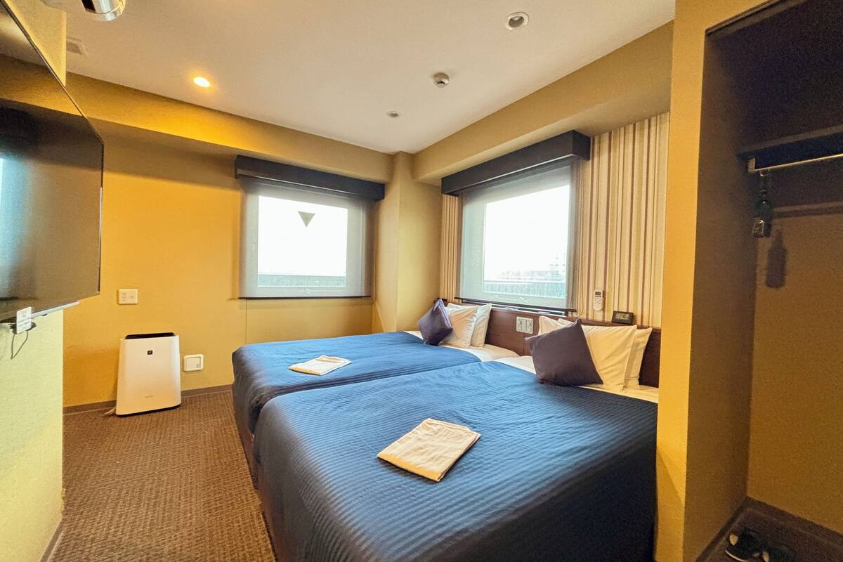 A hotel room with two beds, a small air conditioning unit, a window with curtains, and a door with a handle.