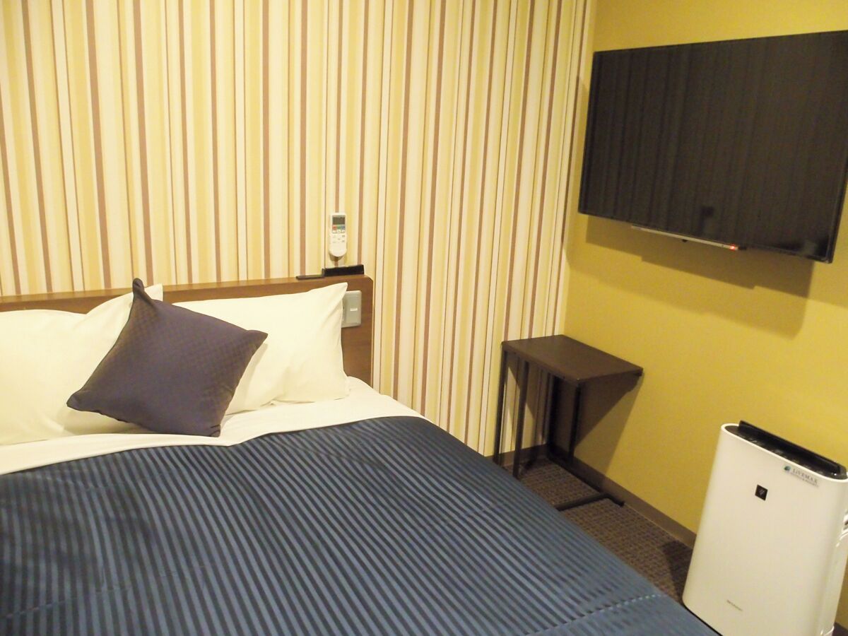 A hotel room with a bed, a bedside table, a TV mounted on the wall, and a white air conditioning unit.